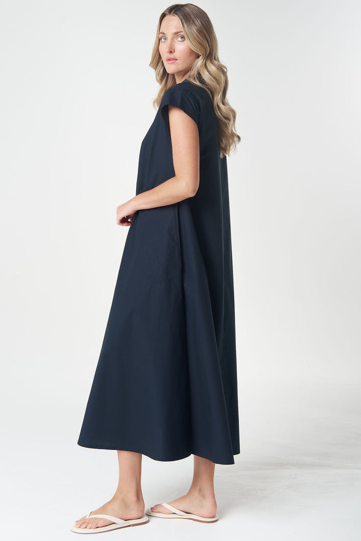 A woman modeling a navy blue midi dress with short cap sleeves and a relaxed, flowing silhouette, paired with white flip-flop sandals, set against a plain light gray studio background.