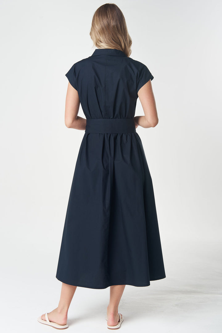 A woman viewed from the back wearing a dark navy midi dress with cap sleeves and a defined waist, paired with white slide sandals, against a light gray studio background.