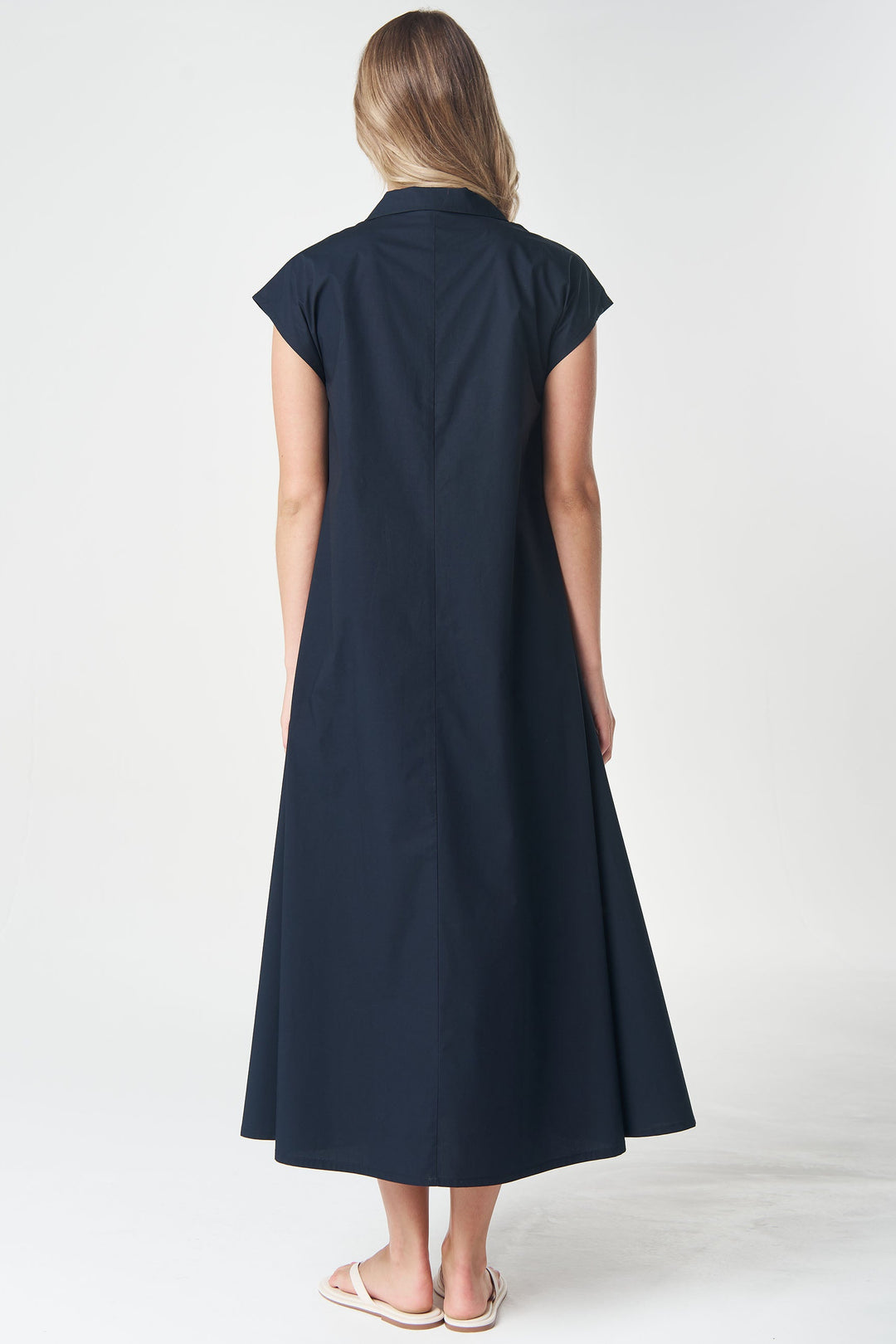 A woman viewed from the back wearing a dark navy midi dress with cap sleeves and a collar, standing against a plain light gray studio background.