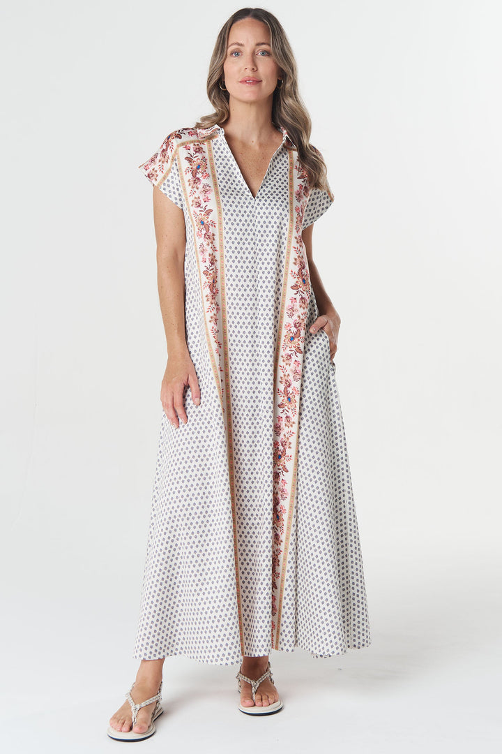 A woman modeling a long white maxi dress with a small red and black polka dot pattern and a pink floral border trim, standing against a plain white studio background.