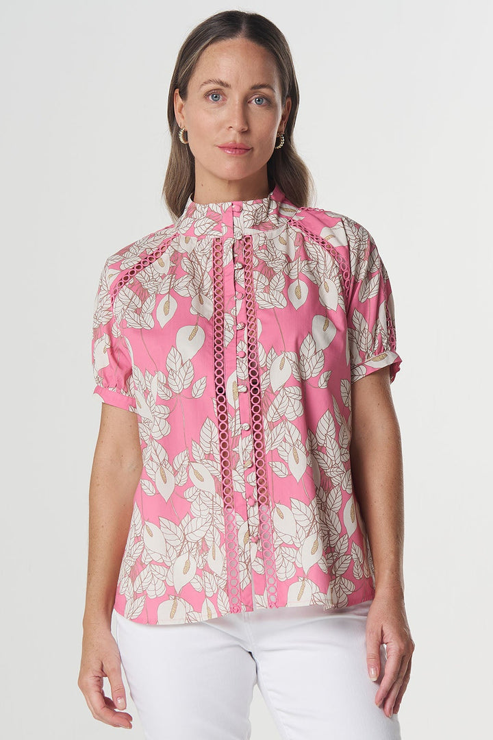 A woman modeling a short-sleeve blouse with a pink and beige floral pattern and a tie-neck detail, paired with white capri pants and heeled sandals, against a light gray studio background.