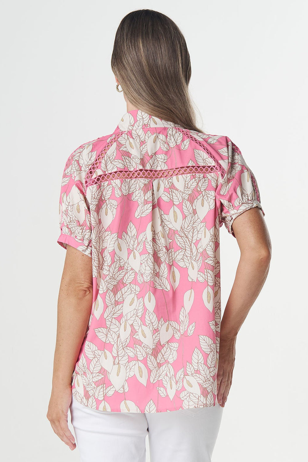 A woman viewed from behind wearing a pink and white floral short-sleeve button-up shirt, white capri pants, and beige heeled sandals against a plain white studio background.