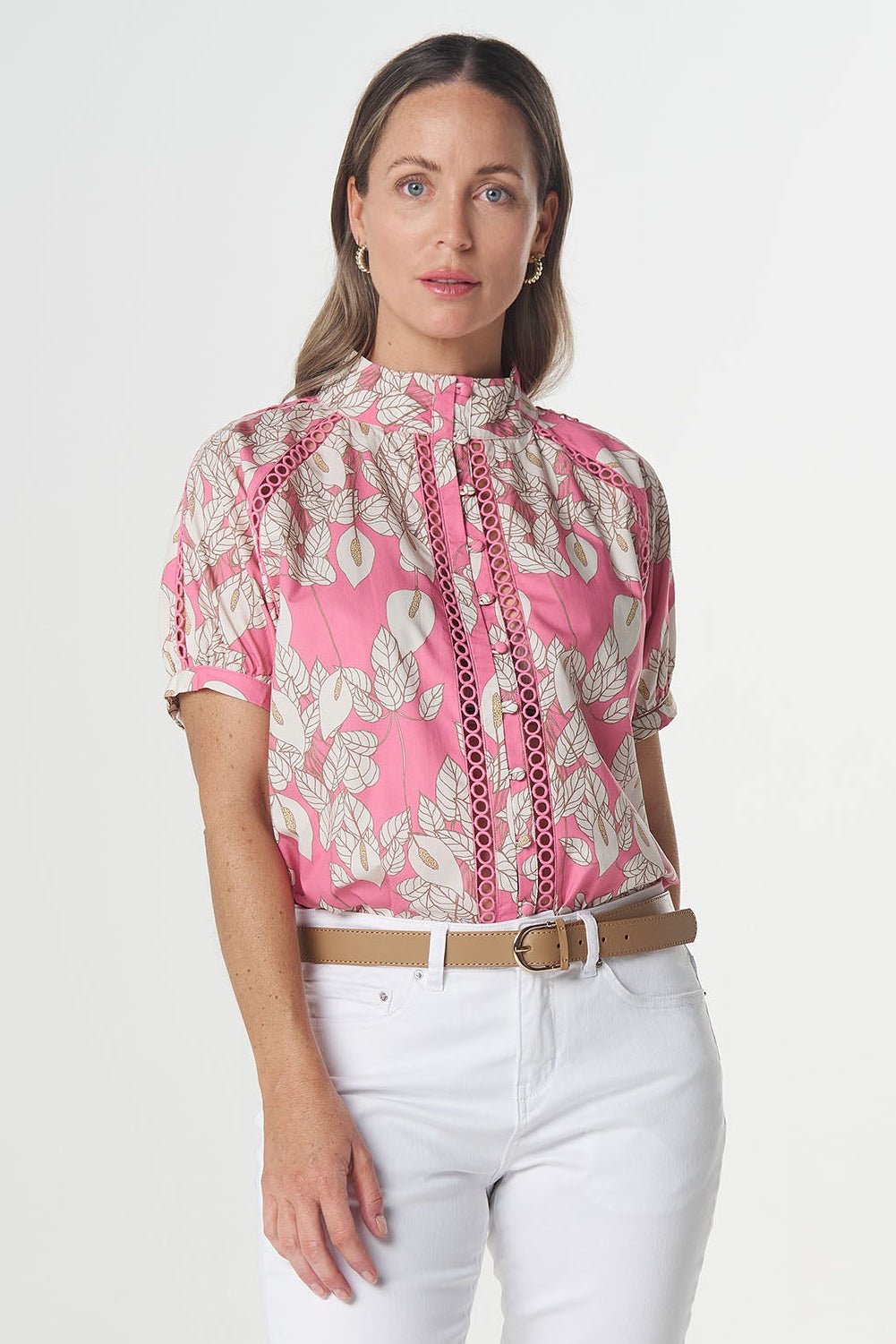 A woman modeling a short-sleeve pink and white patterned blouse with a high gathered neckline, paired with white capri pants and tan sandals, against a plain light gray studio background.
