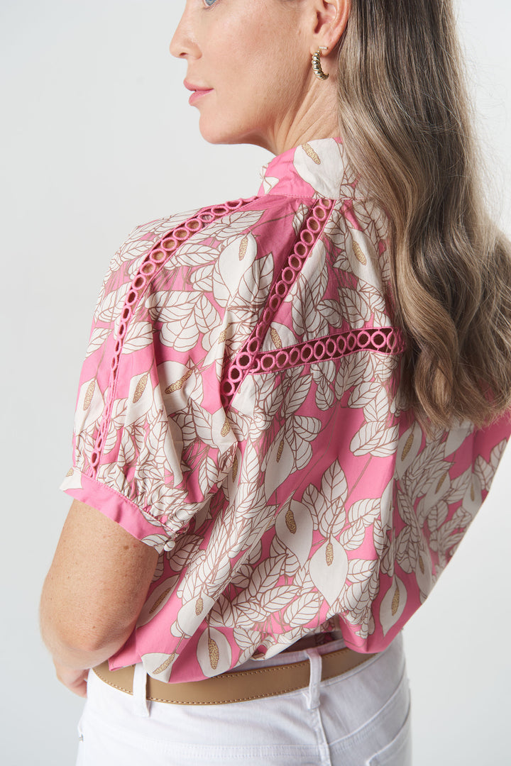 A woman shown from the back wearing a pink and beige floral print blouse with eyelet cutout details along the shoulders, paired with white pants.
