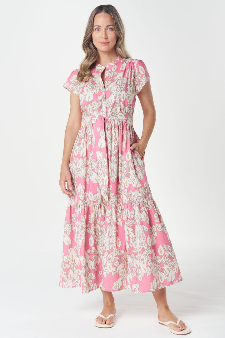 A woman modeling a pink and white floral tiered midi dress with short sleeves and a tie waist, standing against a light gray studio background.