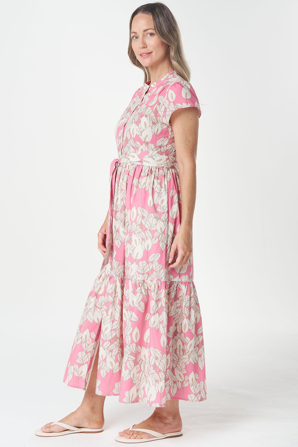 A woman modeling a pink and white patterned midi dress with short sleeves, a tie waist, and a tiered skirt, standing against a plain white studio background.