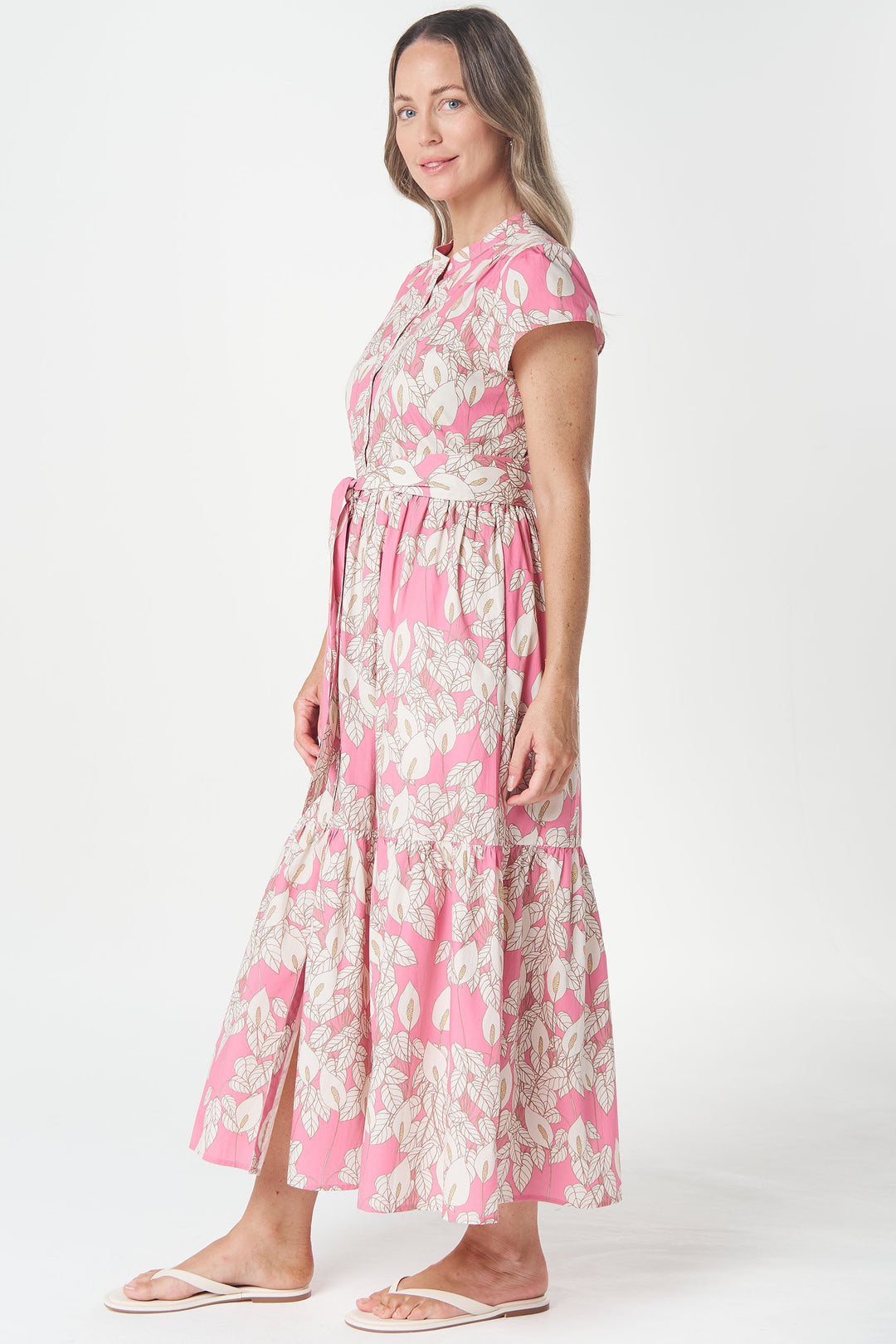 A woman modeling a pink and white patterned midi dress with short sleeves, a tie waist, and a tiered skirt, standing against a plain white studio background.