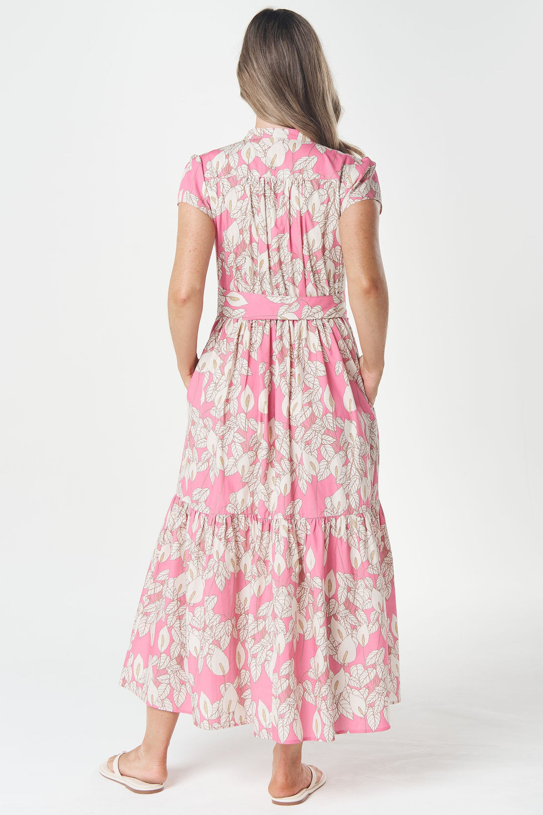 A woman viewed from behind wearing a pink and beige floral tiered midi dress with a matching waist tie, standing against a plain white studio background.