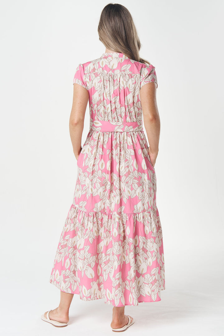 A woman viewed from behind wearing a pink and beige floral tiered midi dress with a matching waist tie, standing against a plain white studio background.