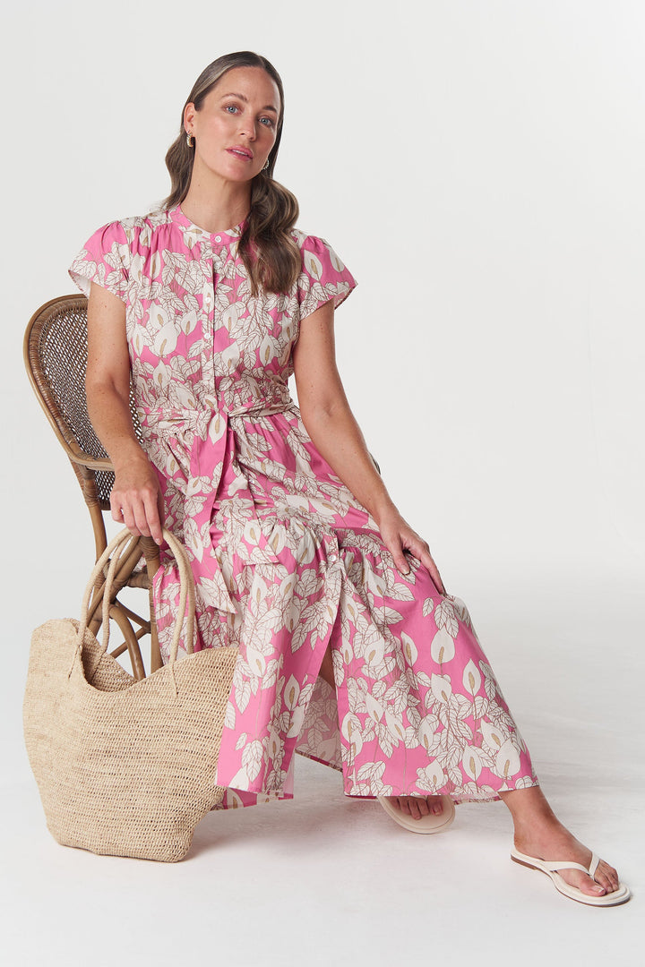 A woman sits on a woven chair wearing a pink and white floral maxi dress, holding a large straw tote bag against a plain light gray studio background.