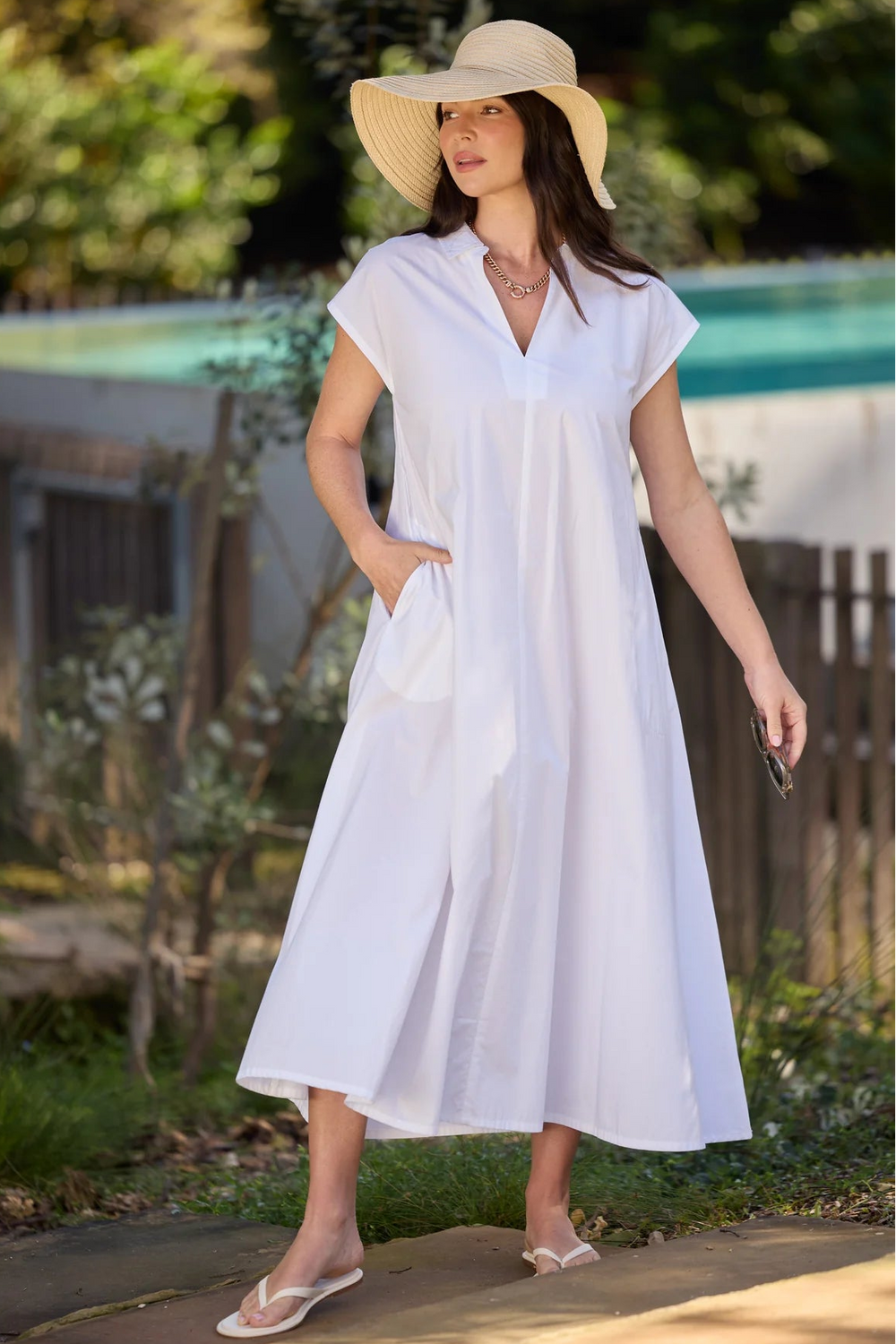 A woman wearing a white midi-length shift dress with short cap sleeves and side pockets, styled with a wide-brimmed straw hat and white flip-flops, standing outdoors near a pool and garden.