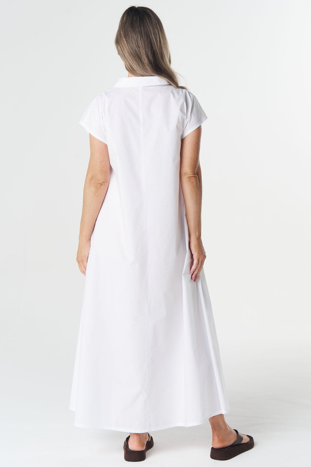 A woman viewed from behind wearing a white short-sleeve collared maxi dress and dark brown sandals, standing against a plain white studio background.