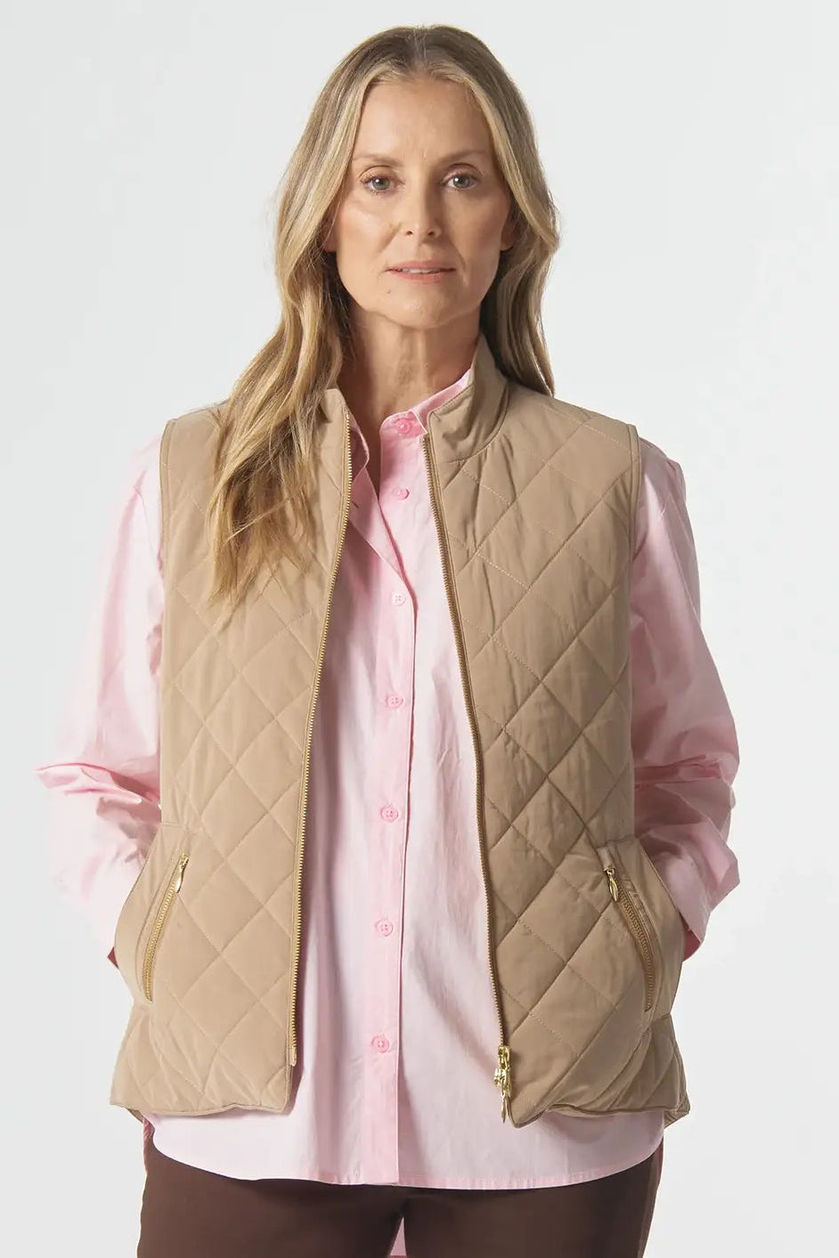 Woman wearing a beige quilted vest, pink shirt, and brown pants on a white background