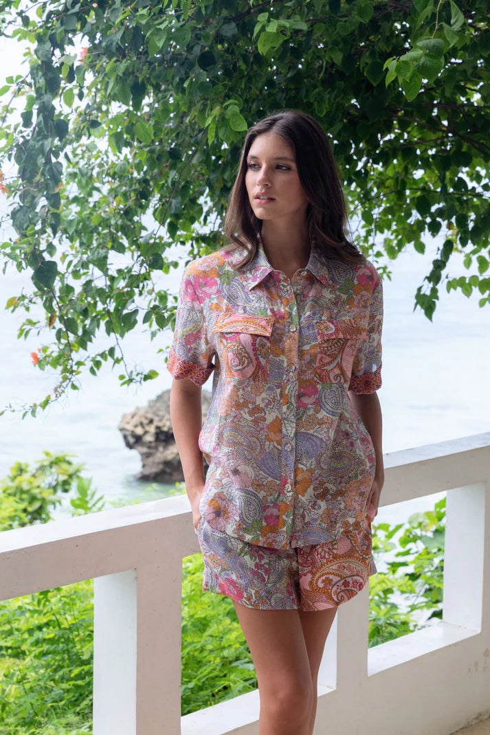 Woman wearing a floral shirt and shorts set on a balcony with greenery and water in the background. Kachel Isla Shirt