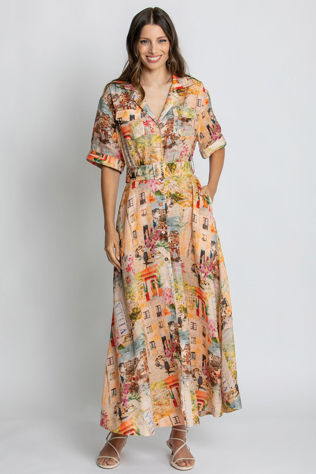 A woman modeling a short-sleeve, collared, V-neck maxi dress with a colorful cityscape print, standing against a light gray studio background.
