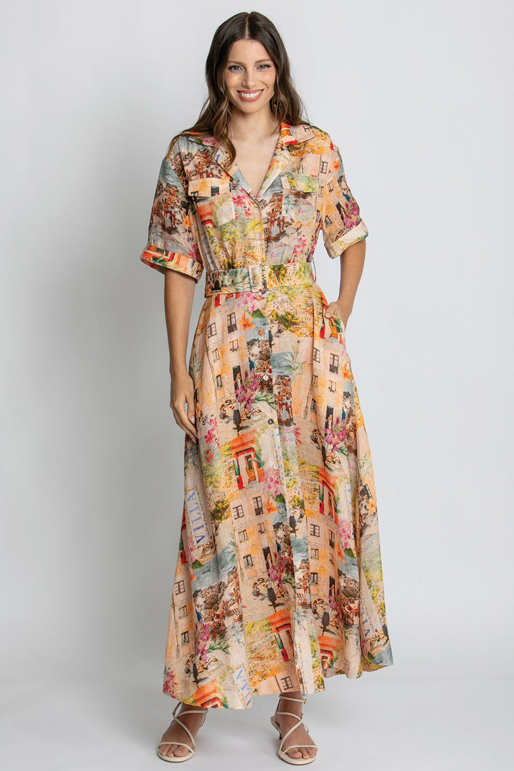 A woman modeling a short-sleeve, collared, V-neck maxi dress with a colorful cityscape print, standing against a light gray studio background.