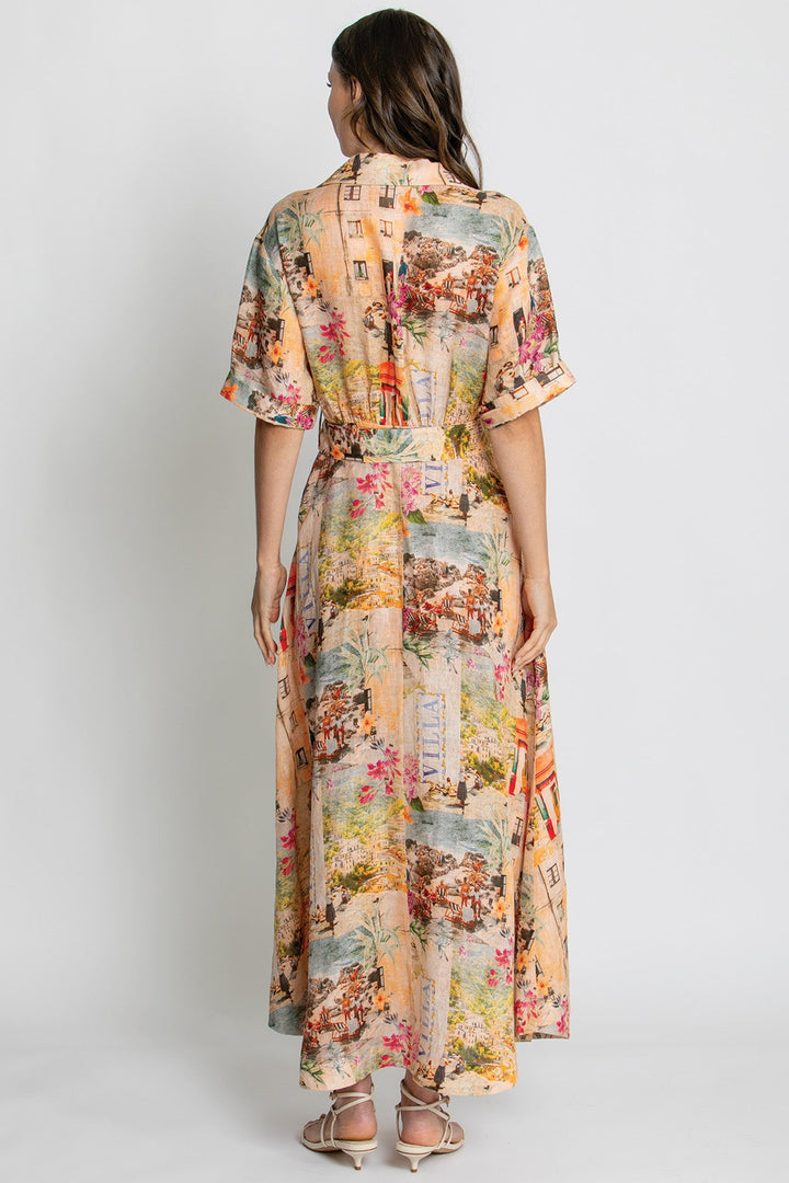 A woman viewed from the back wearing a short-sleeve maxi dress with a multicolor scenic print on a beige base, paired with beige strappy heels, against a light gray studio background.