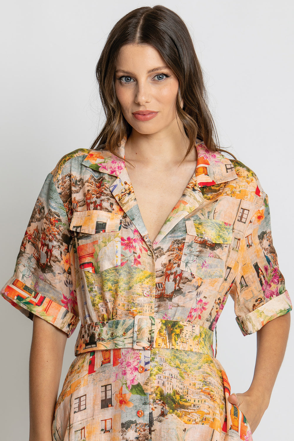 A woman wearing a short-sleeve collared shirt dress with a multicolor cityscape print, featuring a button-up front and a matching tie belt at the waist, set against a plain light gray background.
