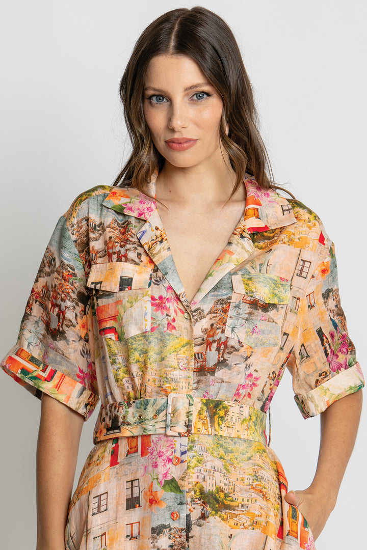 A woman wearing a short-sleeve collared shirt dress with a multicolor cityscape print, featuring a button-up front and a matching tie belt at the waist, set against a plain light gray background.