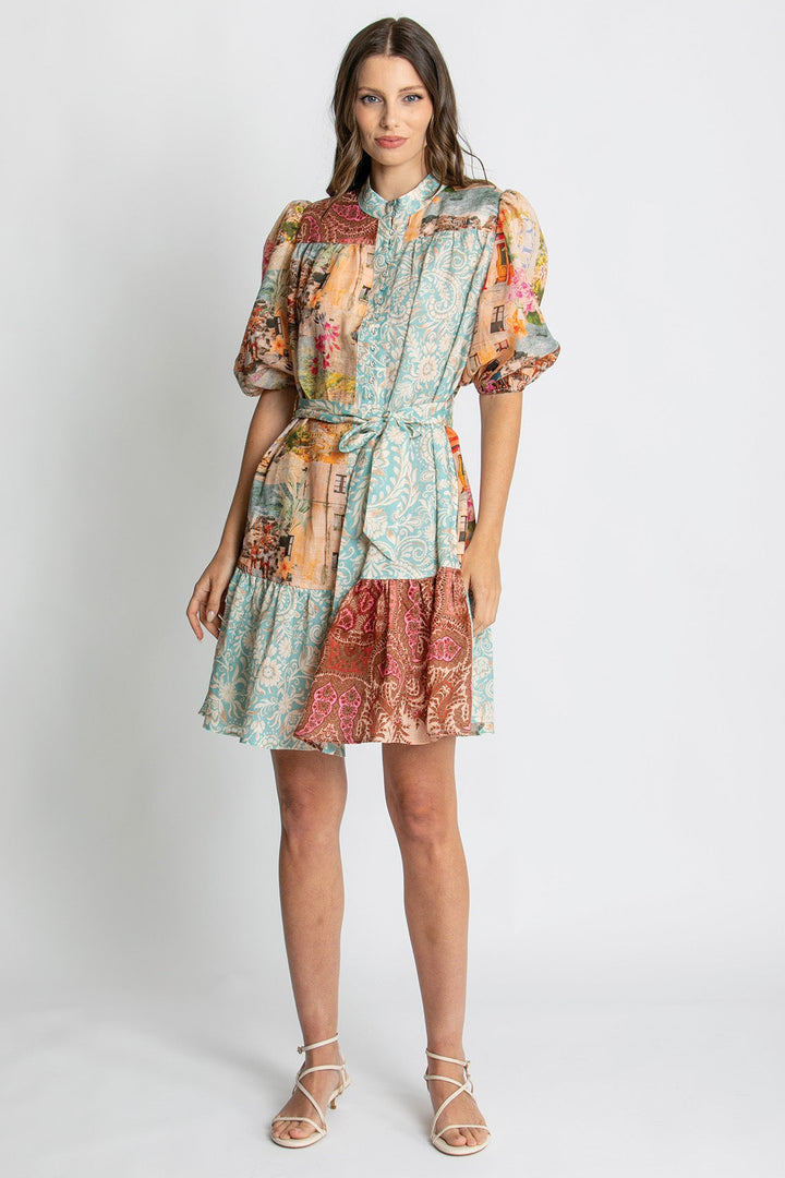 A woman modeling a short puff-sleeve dress with a multicolor patchwork print in shades of teal, rust, beige, and orange, paired with light beige strappy sandals against a light gray studio background.