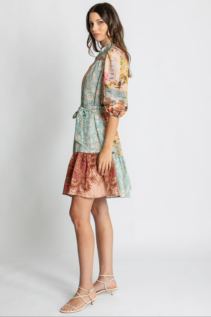A woman modeling a short, multicolor patterned dress with a tie-neck and puff sleeves, paired with beige strappy sandals, against a light gray studio background.