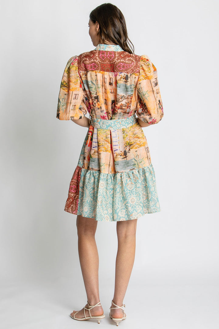 A woman viewed from the back wearing a short puff-sleeve dress with a multicolor patchwork print in orange, beige, red, and light blue, paired with beige strappy heels against a light gray studio background.