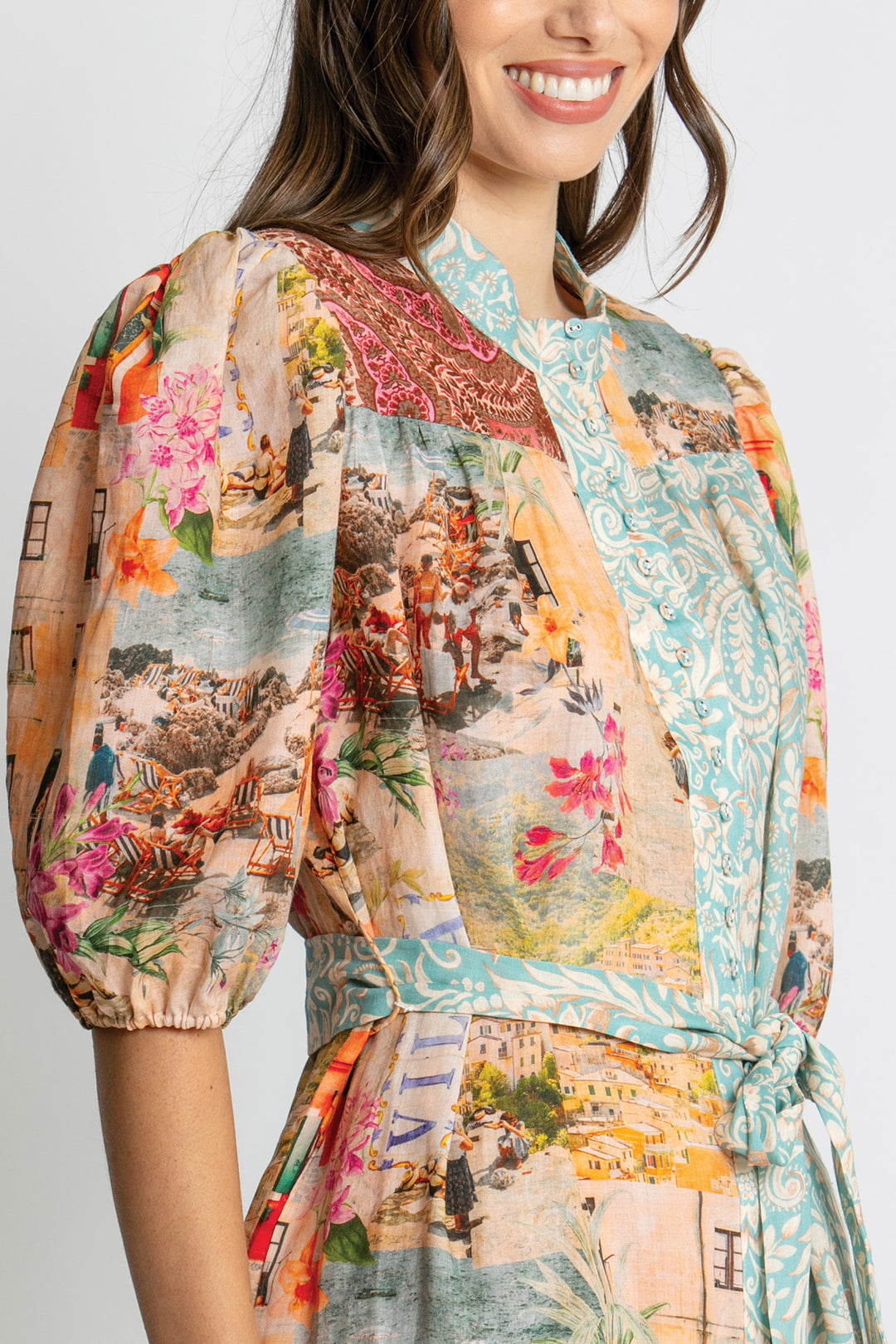 A woman wearing a short puff-sleeve wrap dress with a multicolor collage print featuring landscapes, flowers, and patterns, set against a plain light gray background.