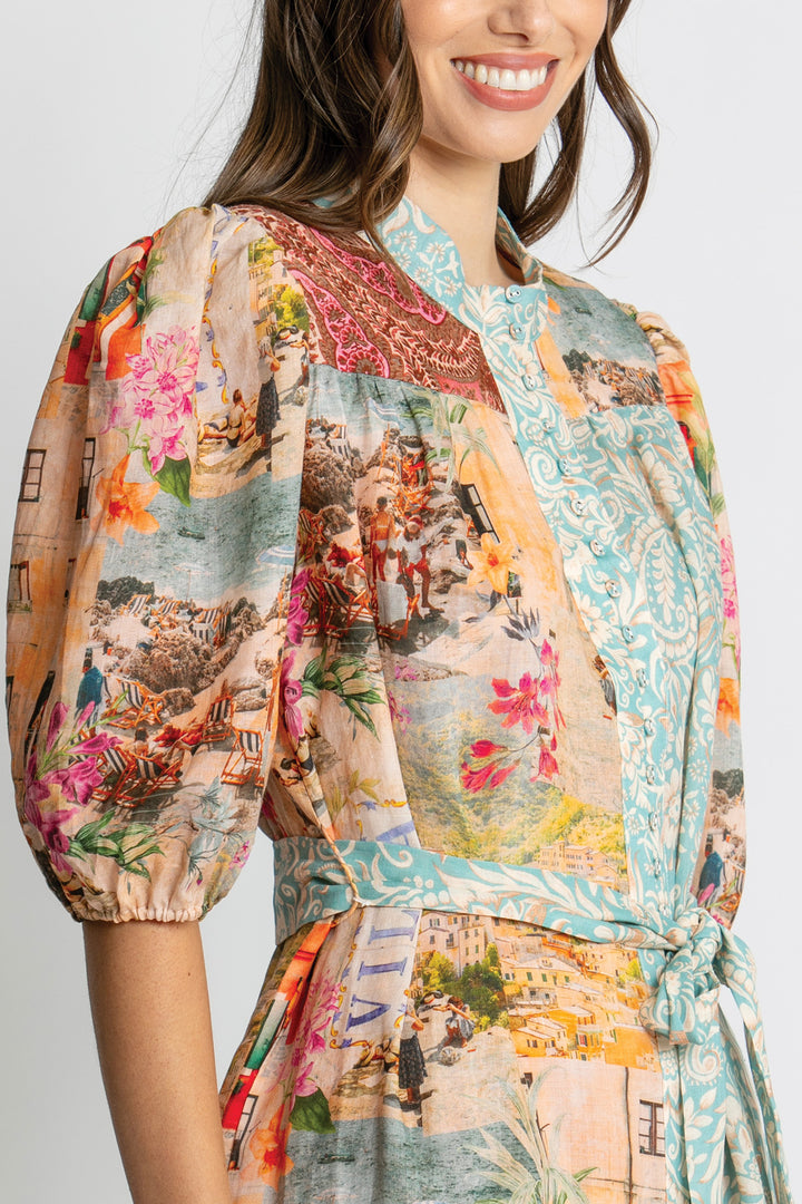 A woman wearing a short puff-sleeve wrap dress with a multicolor collage print featuring landscapes, flowers, and patterns, set against a plain light gray background.