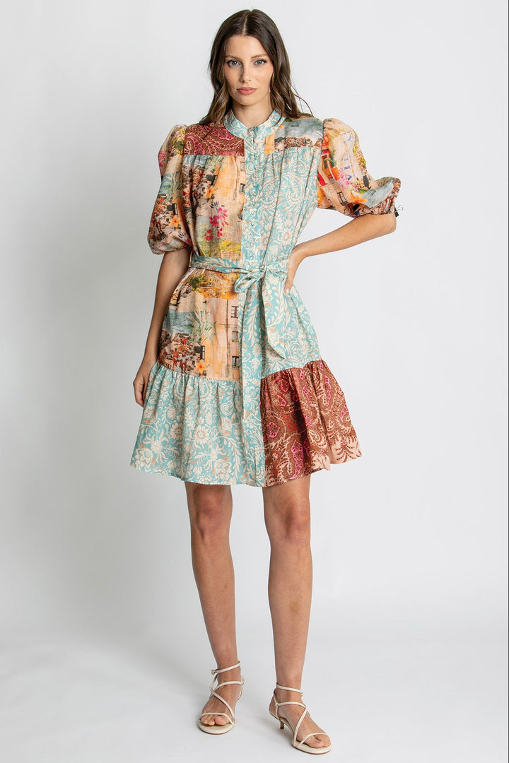 A woman modeling a short patchwork-print dress with puff sleeves, a tie waist, and a tiered skirt in a studio setting.