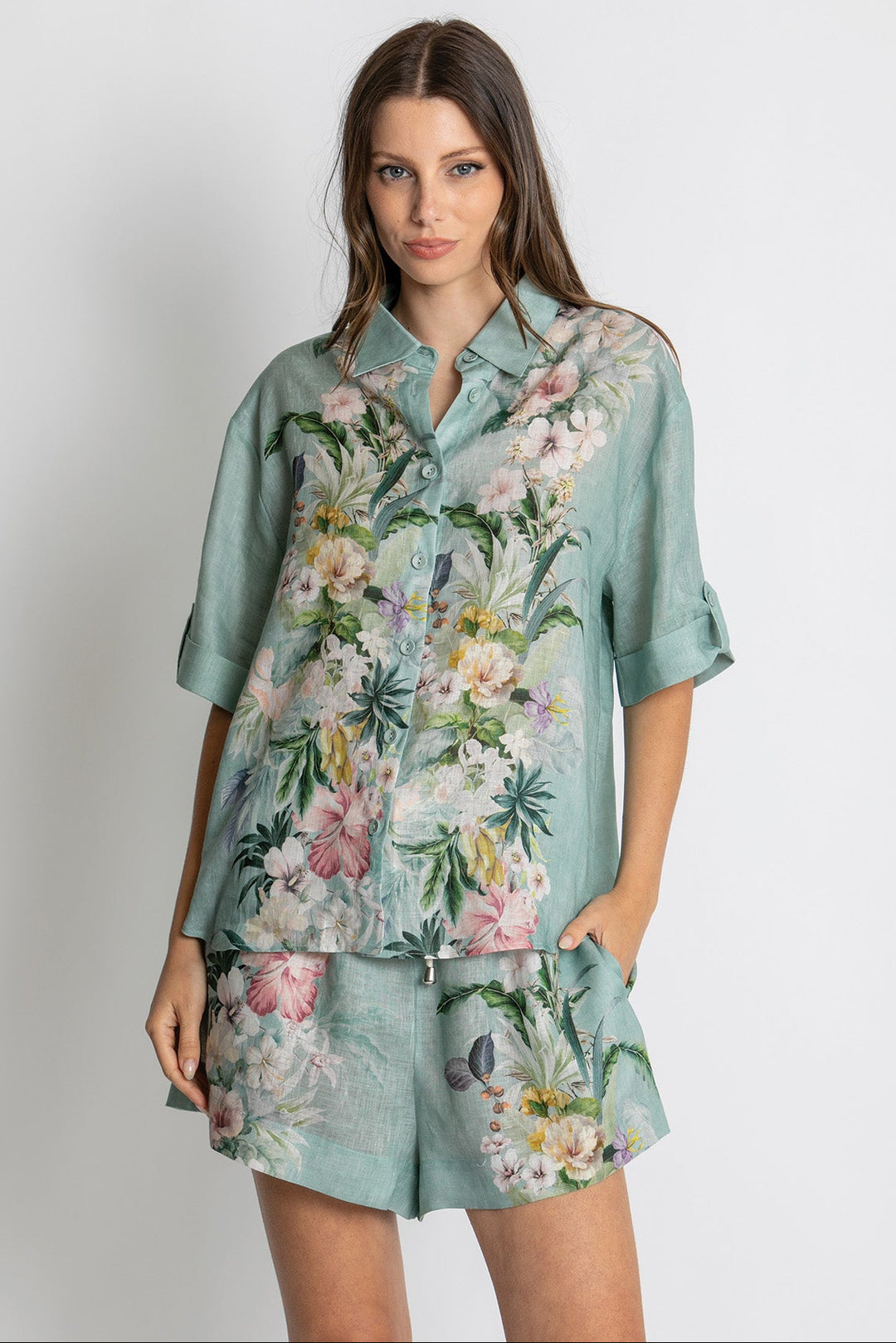 A woman modeling a matching two-piece lounge set consisting of a short-sleeve button-up shirt and shorts in a dusty sage green color with a vibrant pink, white, and green tropical floral print, set against a plain light gray background.