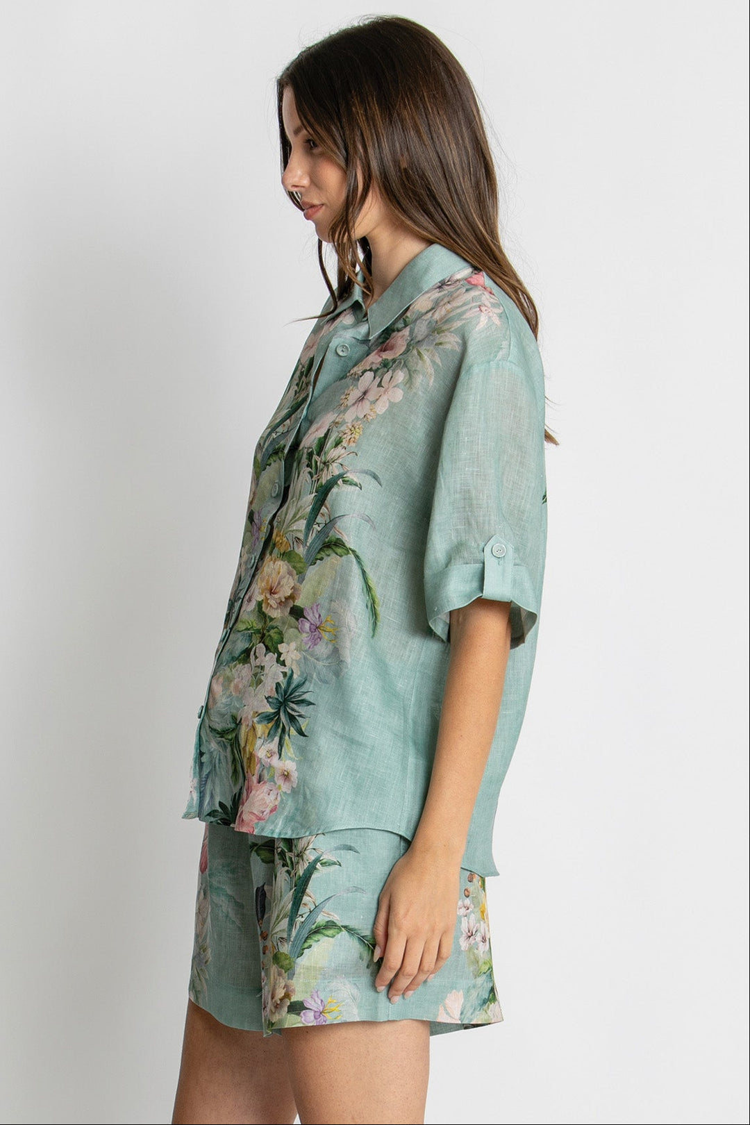 A woman wearing a light blue-green short-sleeve button-up shirt and matching shorts with a pink and green floral print, shown in profile against a plain light gray background.
