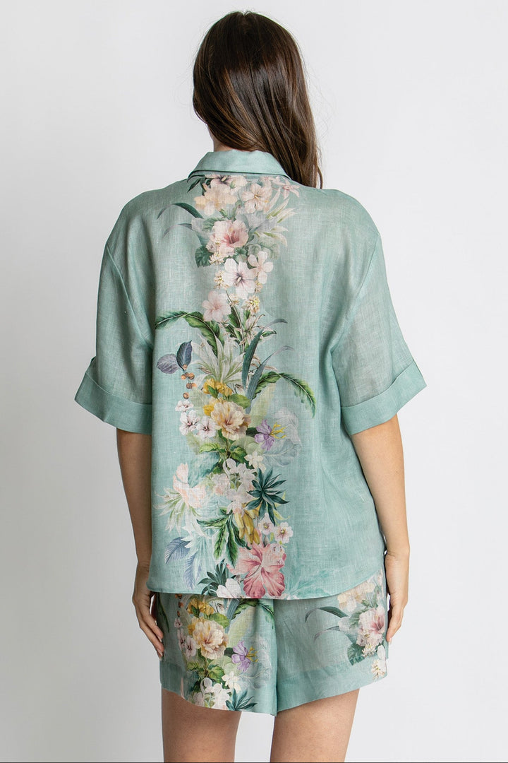 A woman shown from the back wearing a mint green short-sleeve button-up shirt and matching shorts with a large pastel floral print, against a plain light gray background.