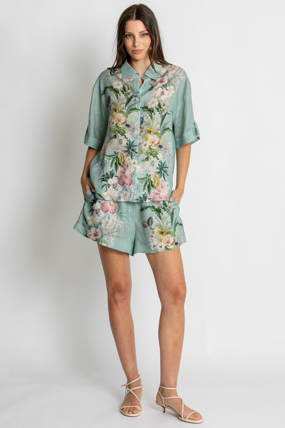 A woman modeling a matching two-piece lounge set featuring a short-sleeve button-up shirt and shorts in a sage green base with a colorful tropical floral print, set against a plain light gray studio background.