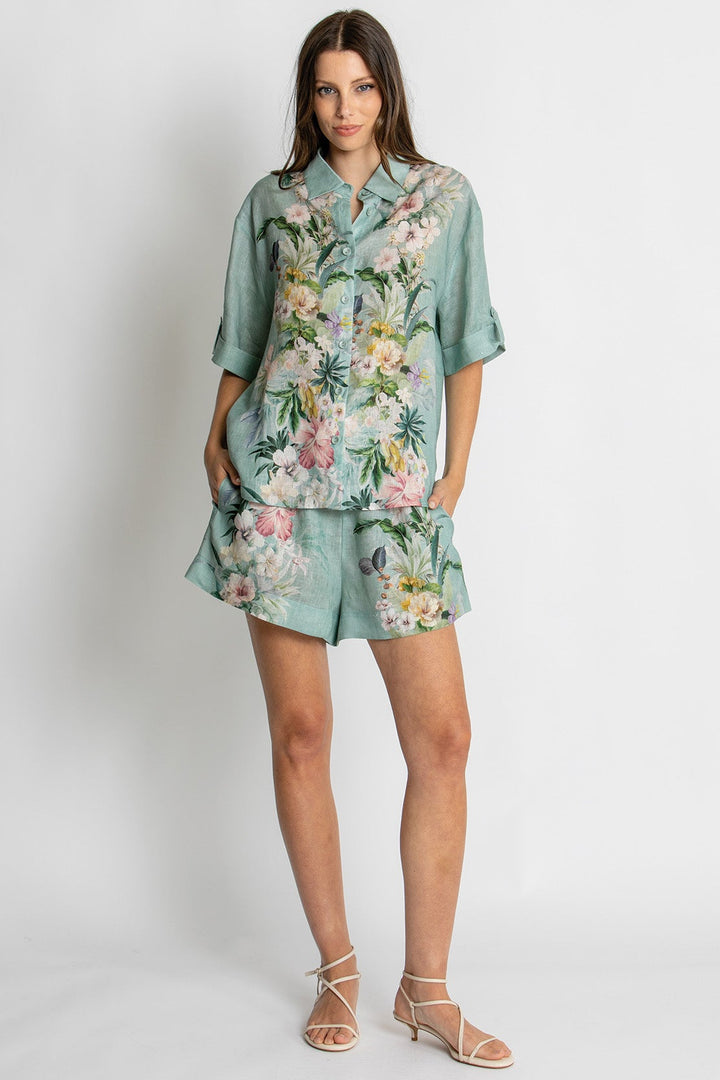 A woman modeling a matching two-piece lounge set featuring a short-sleeve button-up shirt and shorts in a sage green base with a colorful tropical floral print, set against a plain light gray studio background.