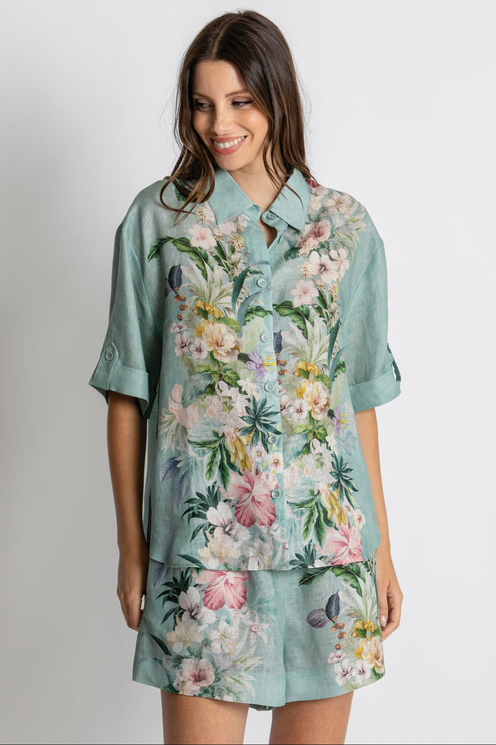 A woman wearing a light teal short-sleeve button-up shirt dress with a colorful tropical floral print, paired with matching shorts, against a plain light gray background.