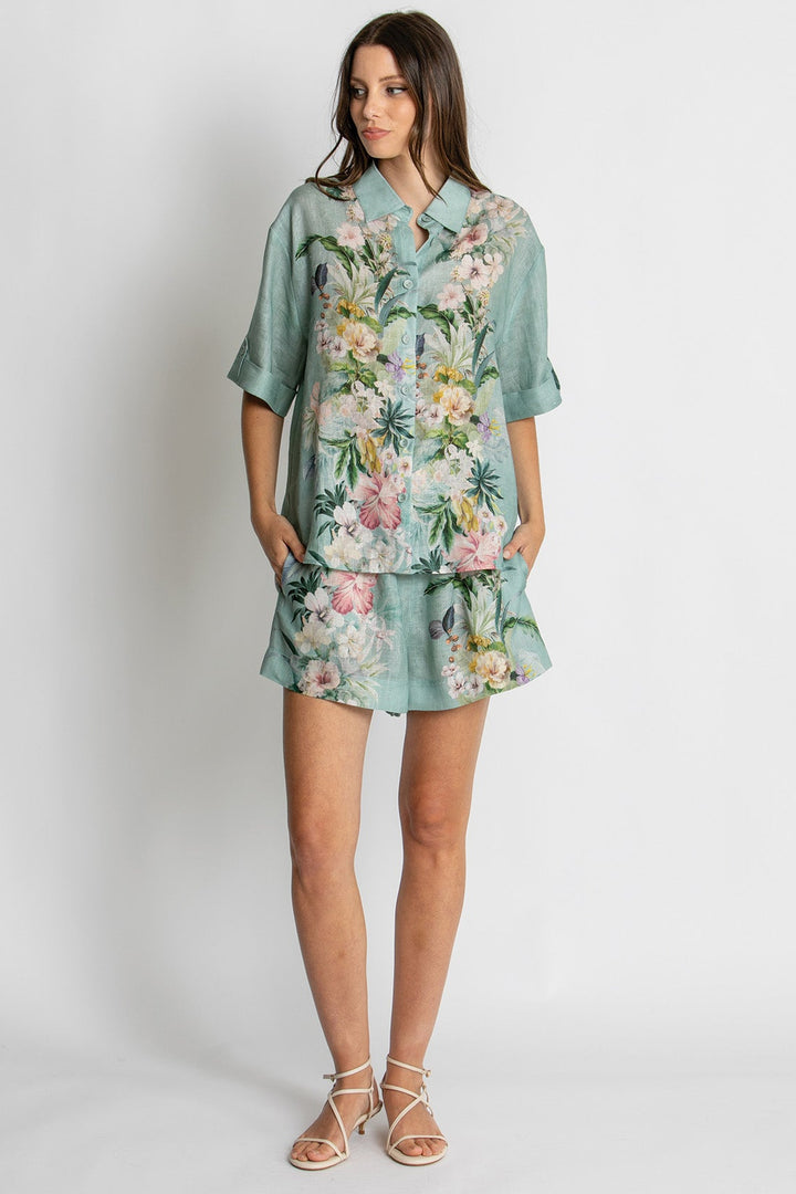 A woman modeling a matching two-piece lounge set featuring a short-sleeve button-up shirt and shorts in a light teal green with a colorful floral print, set against a plain light gray background.