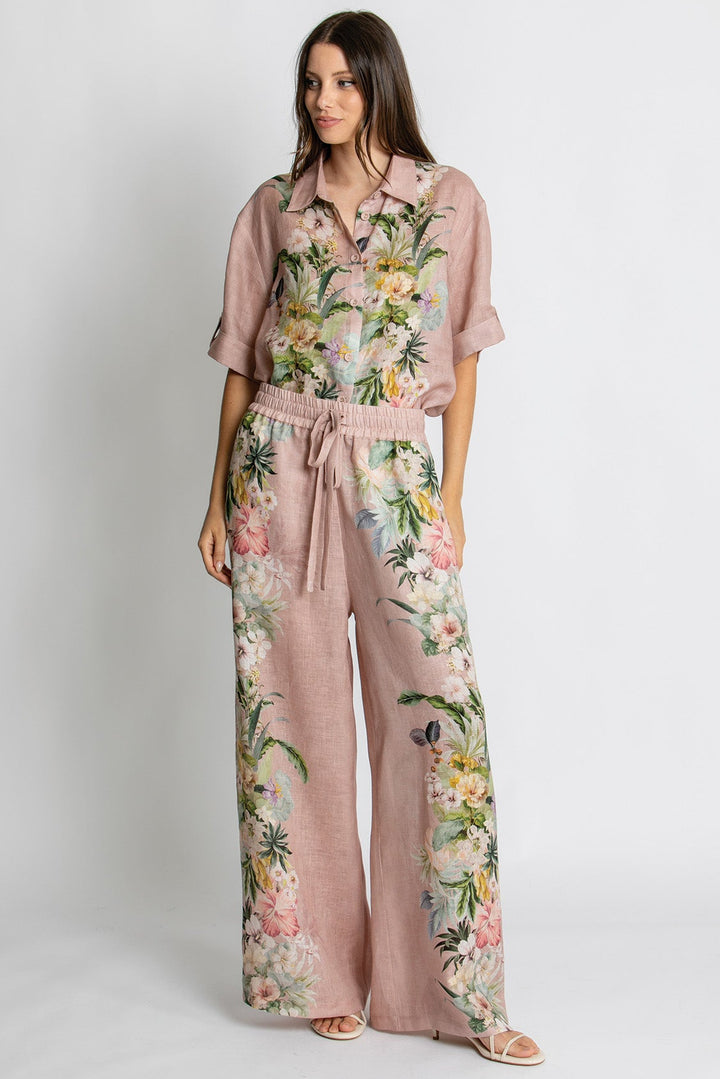 A woman modeling a dusty pink wide-leg jumpsuit with a colorful floral print, featuring a short-sleeve button-up shirt style and a matching drawstring waist, set against a plain light gray studio background.