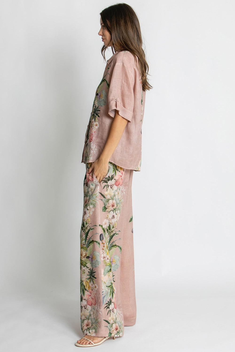 A woman stands in profile wearing a dusty pink two-piece outfit with a short-sleeve button-front top and matching wide-leg pants featuring a colorful floral print.