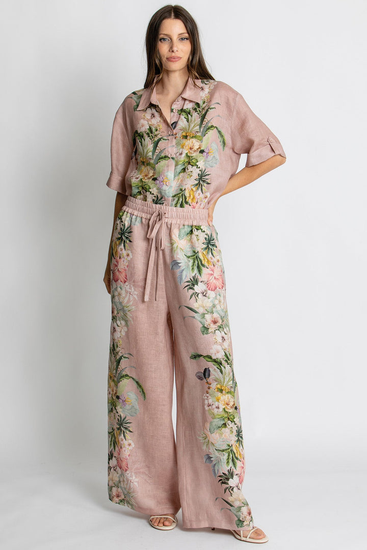 A woman modeling a dusty pink wide-leg jumpsuit with a tropical floral print, featuring short sleeves, a button-up collar, and a matching fabric tie belt at the waist, set against a plain light gray studio background.
