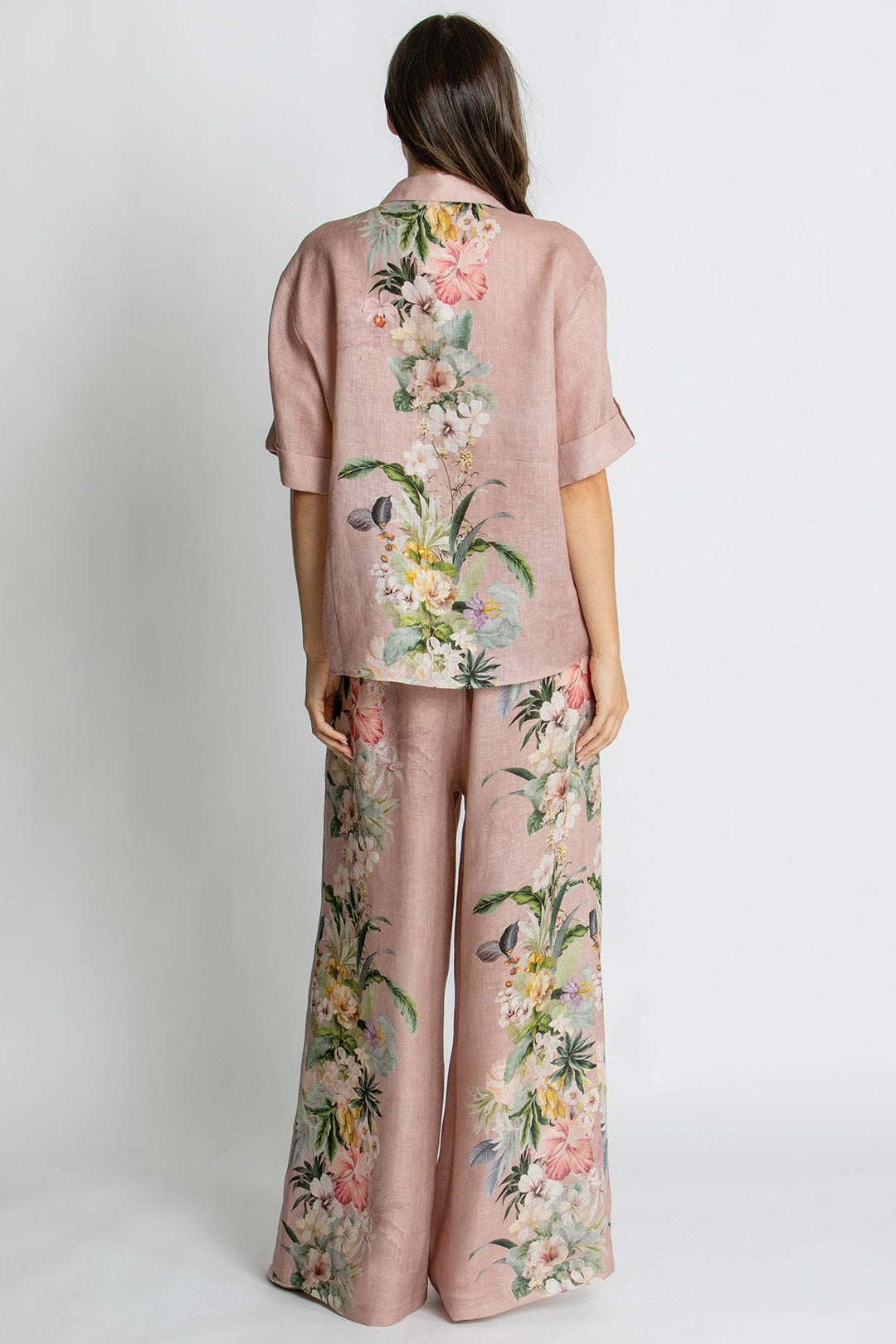 A model viewed from the back wearing a matching two-piece set consisting of a short-sleeve button-up shirt and wide-leg trousers, both in a dusty rose pink color with a vibrant multicolor floral print.