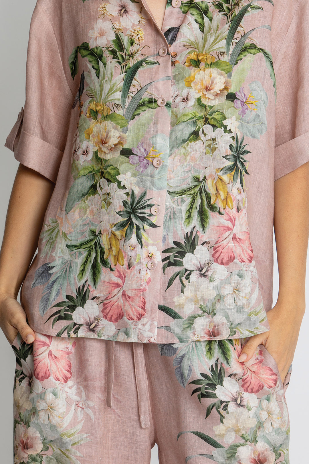 A close-up of a person wearing a matching two-piece pajama set in dusty pink with a large tropical floral print in pink, green, white, and yellow.