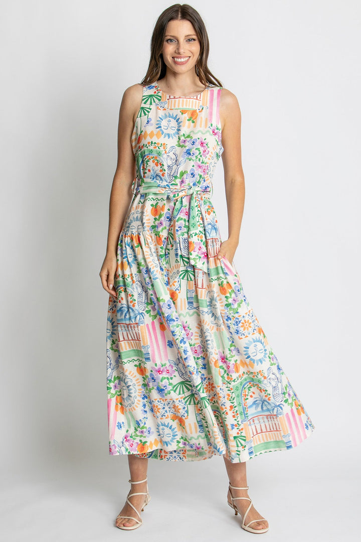 A woman modeling a sleeveless midi dress with a colorful tropical and architectural print, standing against a light gray studio background.