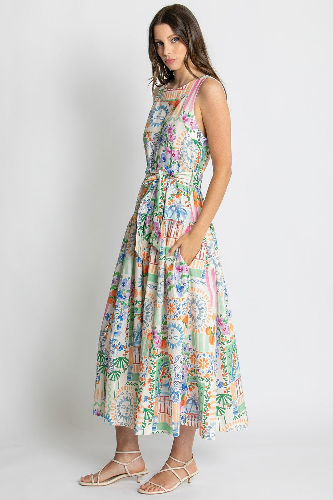 A woman modeling a sleeveless midi dress with a colorful tropical and architectural print, paired with beige strappy sandals, set against a plain light gray studio background.