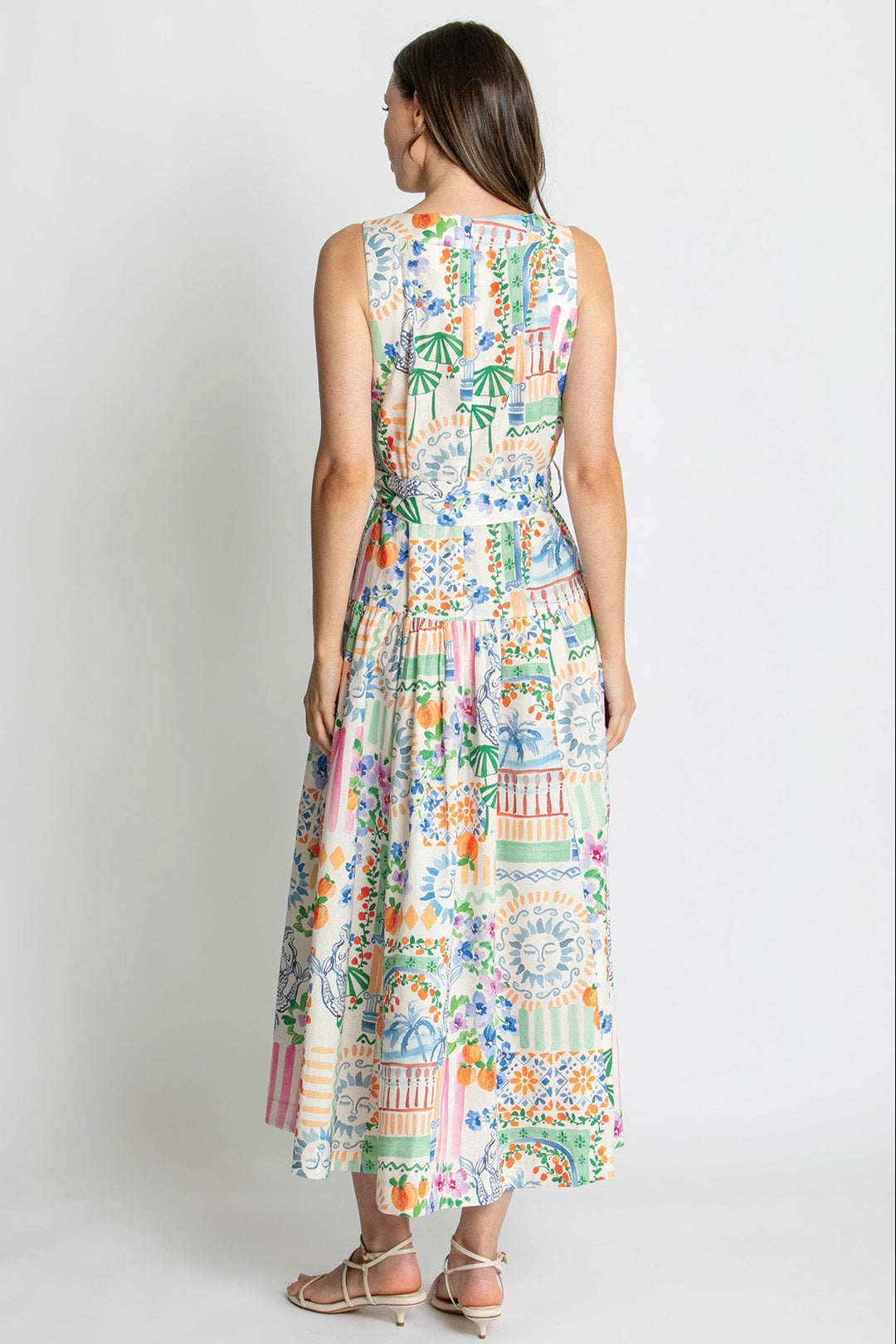A woman viewed from the back wearing a sleeveless, knee-length maxi dress with a colorful patchwork tropical and floral print, paired with beige strappy high-heeled sandals, against a light gray studio background.