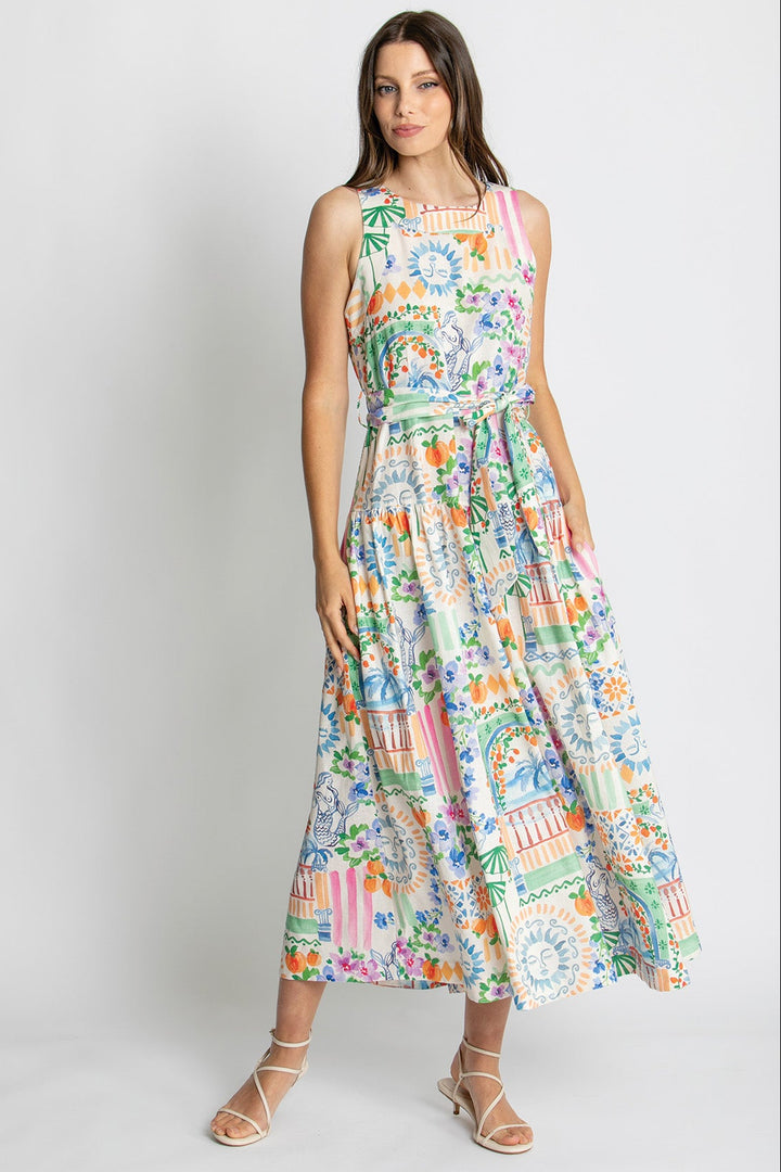 A woman modeling a sleeveless, high-neck, midi-length maxi dress with a vibrant multicolor tropical and architectural print, paired with beige strappy sandals against a light gray studio background.