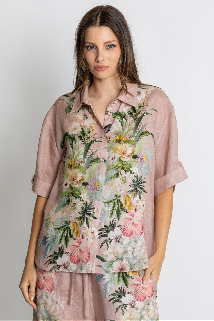 A woman modeling a dusty pink short-sleeve button-up shirt with a vibrant tropical floral print in green, pink, and white, paired with matching floral pants against a light gray studio background.