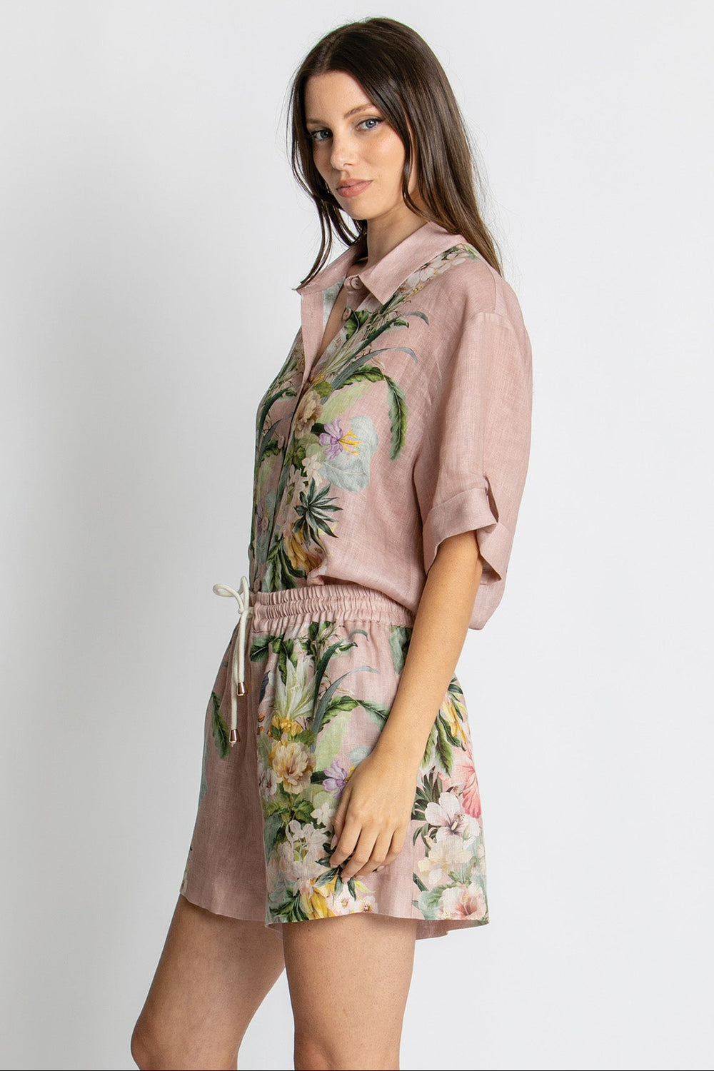 A woman modeling a dusty pink short-sleeve collared shirt and matching drawstring shorts set with a tropical floral print, set against a plain light gray background.