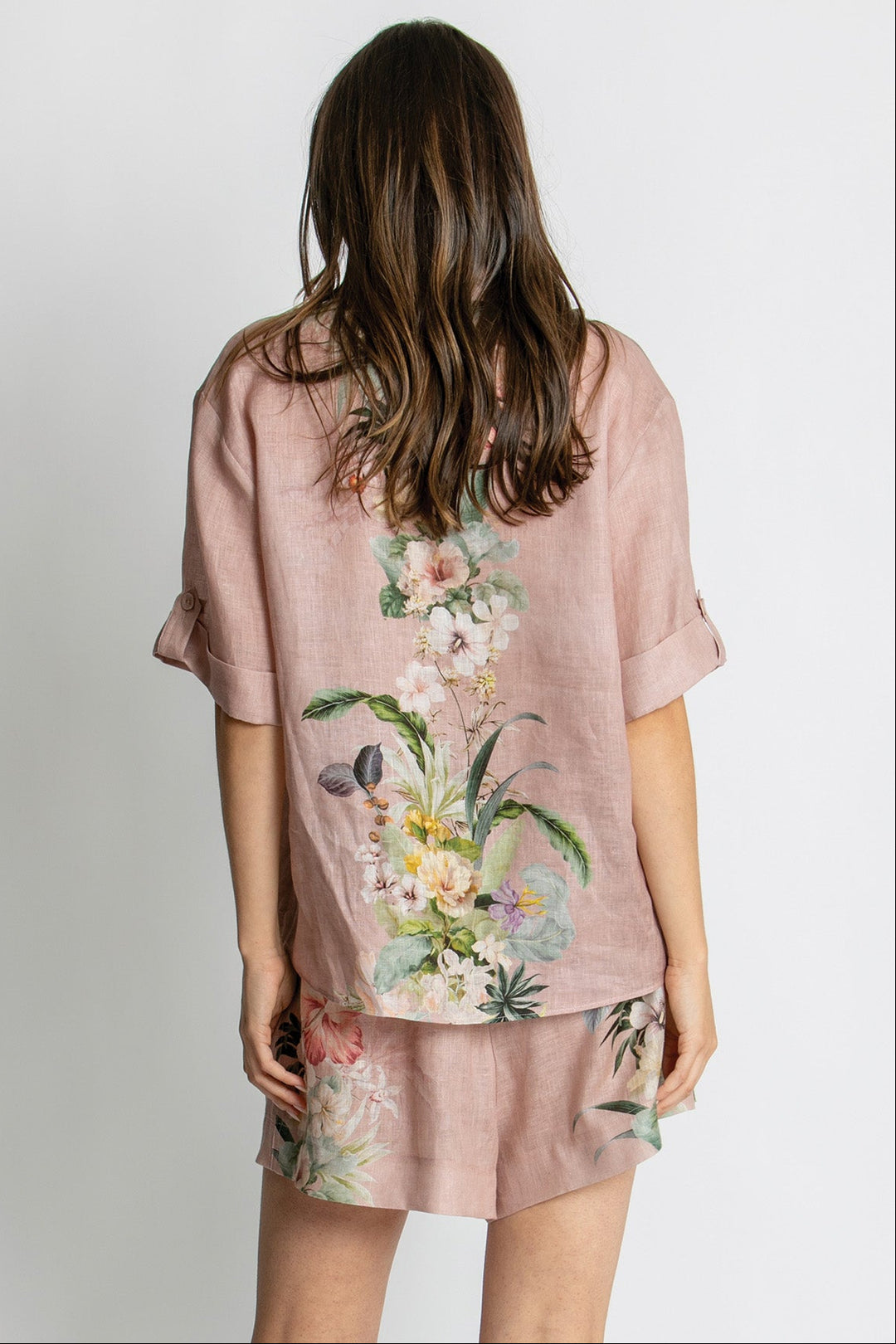 A woman viewed from the back wearing a dusty pink short-sleeve top and matching shorts set with a large floral print on the back and upper thighs, against a plain light gray background.