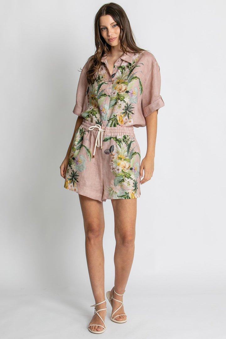 A woman modeling a dusty pink short-sleeve button-up shirt and matching shorts set with a tropical floral print, paired with beige strappy high-heeled sandals, against a light gray studio background.