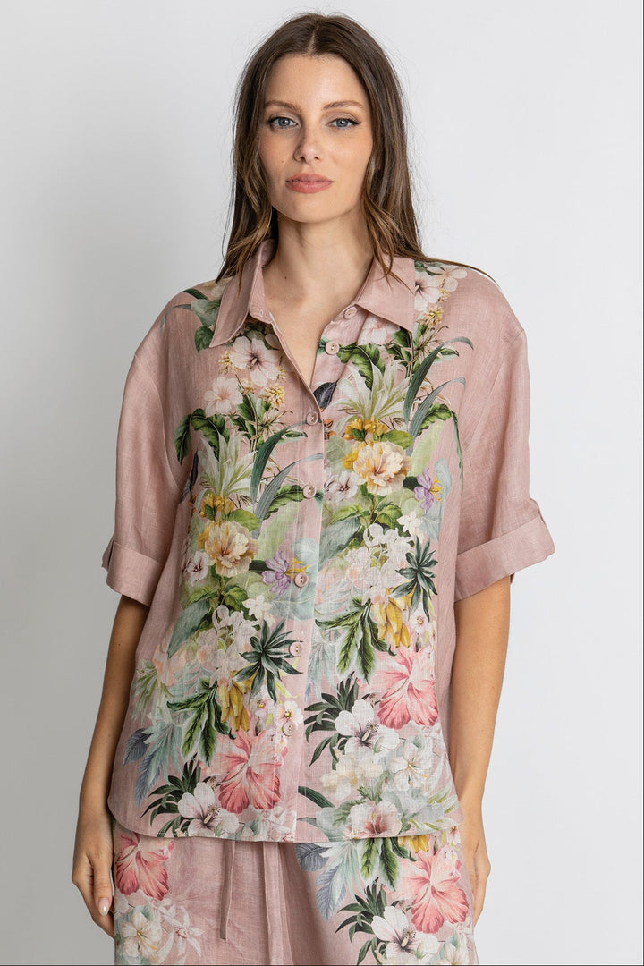 A woman modeling a dusty pink short-sleeve button-up shirt with a vibrant tropical floral print in green, pink, and white, paired with matching floral pants against a light gray studio background.